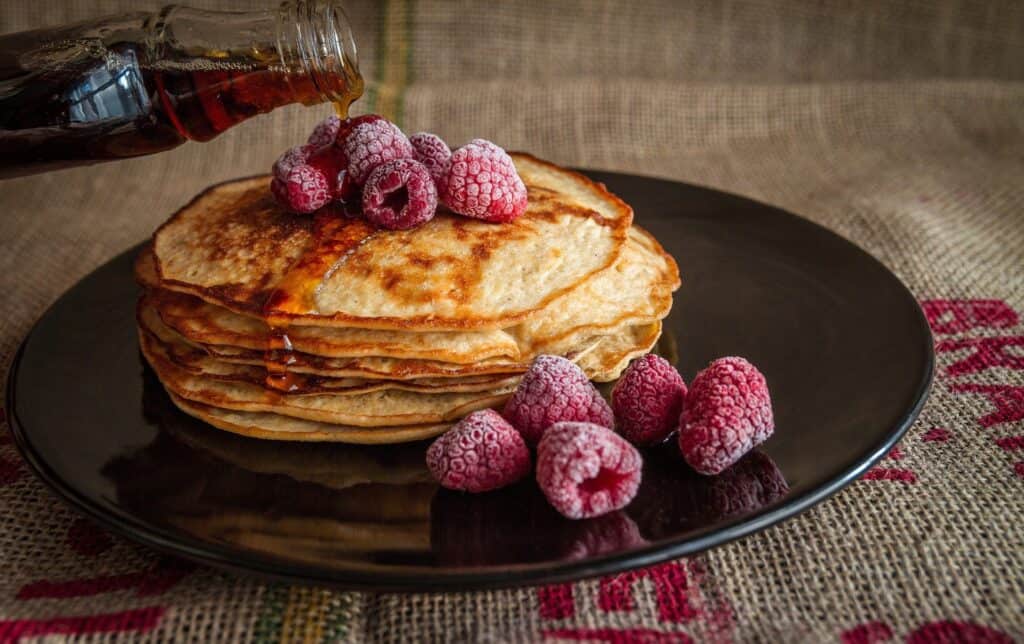 Maple syrup being poured on a stack of pancakes topped with frozen raspberries on a black plate