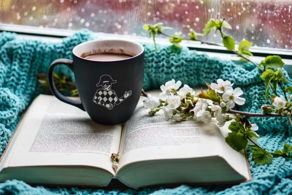 Mug of hot chocolate on top of a book surrounded by teal knitwear and white flowers on branches