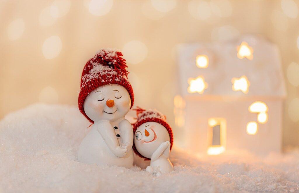 Title image for Fall & Winter Energy Saving Tips - two model snowmen in red woollen hats standing on fake snow in front of a lit white model house